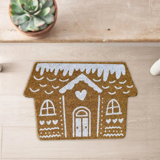 Gingerbread house-shaped doormat on a wooden floor with a plant in the background